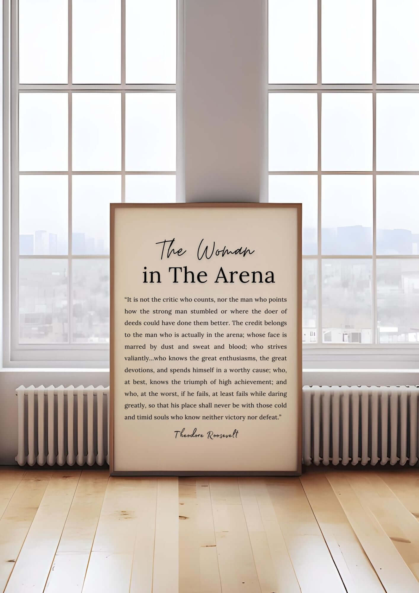 Framed 'The Woman in The Arena Quote' by Theodore Roosevelt on wooden floor in front of large windows.