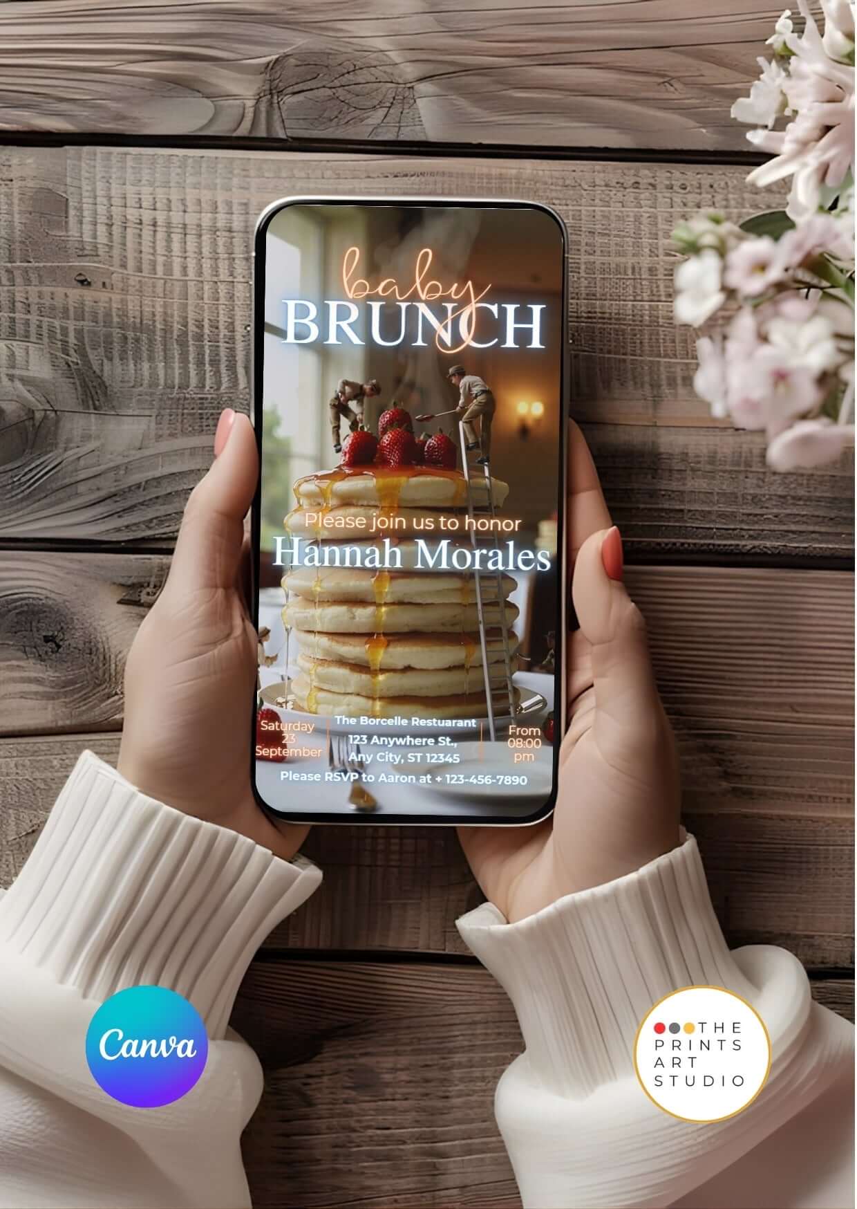 Hands holding a phone displaying a Baby Shower Video Invitation Dreamy Canva Editable with a brunch theme on a wooden background.
